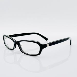 May include: Black-framed spectacles with a rectangular shape and clear lenses. The glasses have a sleek design with a silver accent on the temples. The frames are thick and the arms curve slightly. The glasses are set against a white background.