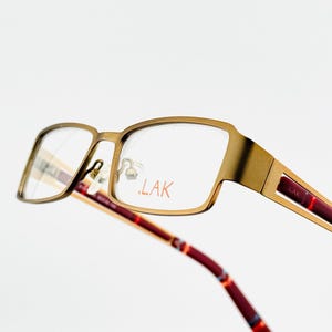 May include: Close-up of eyeglasses with a gold-coloured frame and clear lenses. The temples are burgundy with a colourful pattern. The brand name "LAK" is printed in red on the lens and temple.
