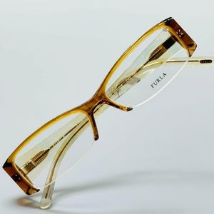 Puede incluir: Gafas Furla con montura marrón claro y carey. Las gafas tienen forma rectangular y lentes transparentes. El logotipo de Furla es visible en la lente. Las patillas son de color beige claro.