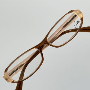 Puede incluir: Gafas con montura marrón con borde y patillas de color melocotón claro. Las lentes son transparentes y se ve un pequeño logotipo de pájaro en la lente superior izquierda. Las gafas están en ángulo sobre un fondo blanco.