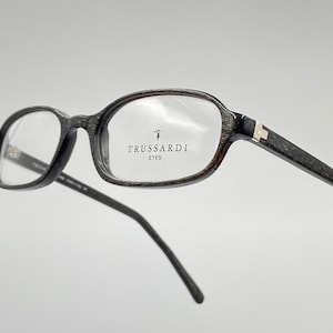 May include: Black eyeglasses with a textured frame and clear lenses. The brand name "TRUSSARDI EYES" is printed on the right lens.