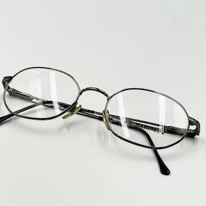 May include: Silver-toned metal eyeglasses with oval-shaped lenses. The glasses have a black temple arm and the text "G. MARINES" is printed on the right lens.