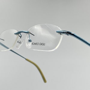 May include: Rimless eyeglasses with blue metal frames and temples. The lenses are clear, and the nose pads are transparent. The brand name "ROMEO GIGLI" is printed on the right lens. The temples have a light tan color at the ends.