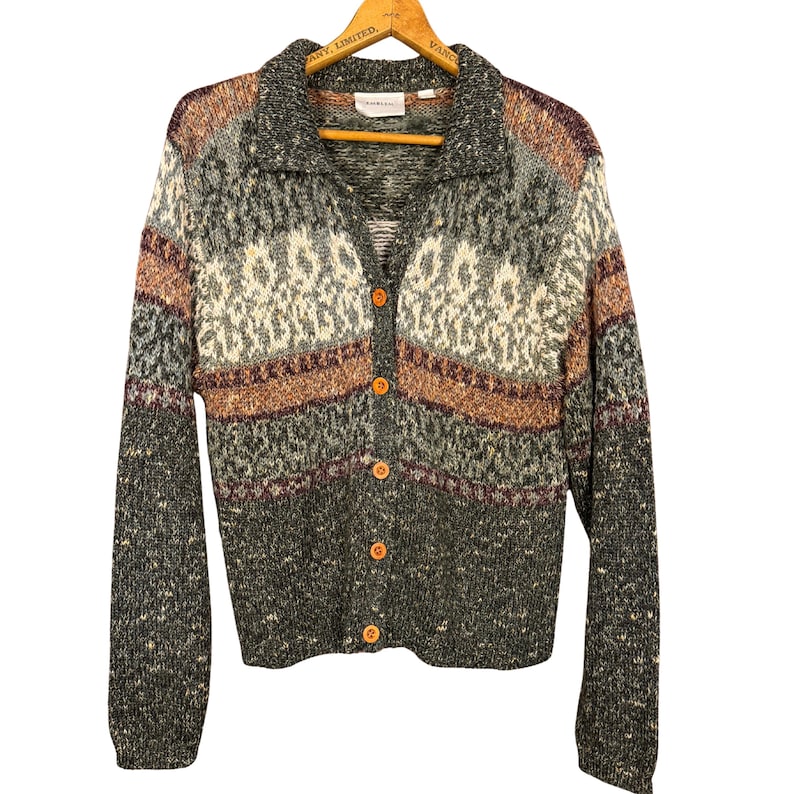 May include: A button-down cardigan sweater with a collared neckline. The sweater features a pattern of gray, brown, cream, and burgundy. The buttons are a light orange color. The sweater is hanging on a wooden hanger.