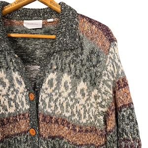 May include: A button-down cardigan sweater in shades of green, gray, brown, and cream. The sweater has a collared neckline and two orange buttons. The brand name "EMBLEM" is visible on a tag near the collar. The sweater is hanging on a wooden hanger.