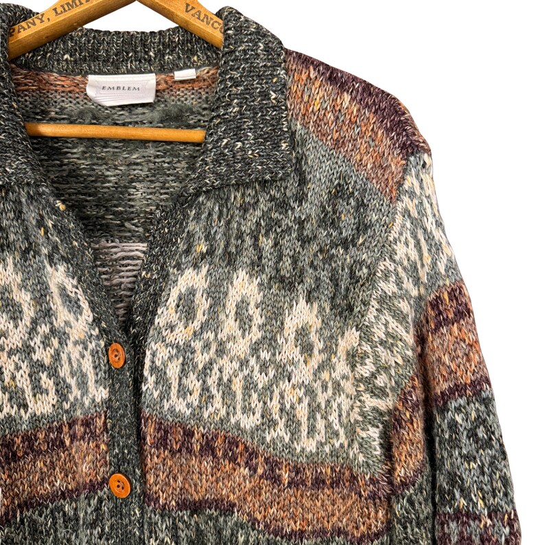 May include: A button-down cardigan sweater in shades of green, gray, brown, and cream. The sweater has a collared neckline and two orange buttons. The brand name "EMBLEM" is visible on a tag near the collar. The sweater is hanging on a wooden hanger.