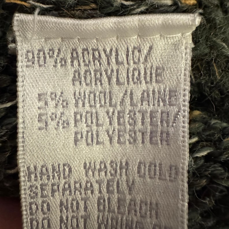 May include: A close-up of a white fabric tag with black text. The tag lists the material composition as 90% acrylic, 5% wool, and 5% polyester. Washing instructions include hand wash cold and do not bleach.
