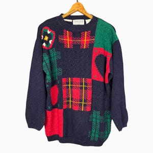 Könnte beinhalten: Ein marineblauer Pullover mit einem Patchwork-Design mit roten, grünen und gelben Karomustern und Blumenmustern. Der Pullover hat einen Rundhalsausschnitt und lange Ärmel. Der Pullover hängt an einem Holzkleiderbügel.