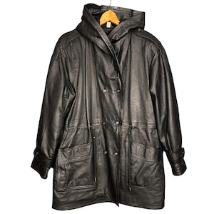 Op de afbeelding: Zwarte leren jas met capuchon, dubbele rij knopen, twee grote voorzakken en verstelbare taille. De jas heeft een taille met trekkoord en manchetten met knopen. De jas hangt aan een houten kleerhanger.