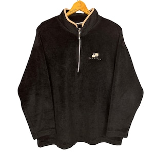 May include: Black fleece pullover with a beige-lined collar and a silver zipper. The pullover features the text "SAN DIEGO" embroidered in white, along with a small logo. The garment is displayed on a wooden hanger.