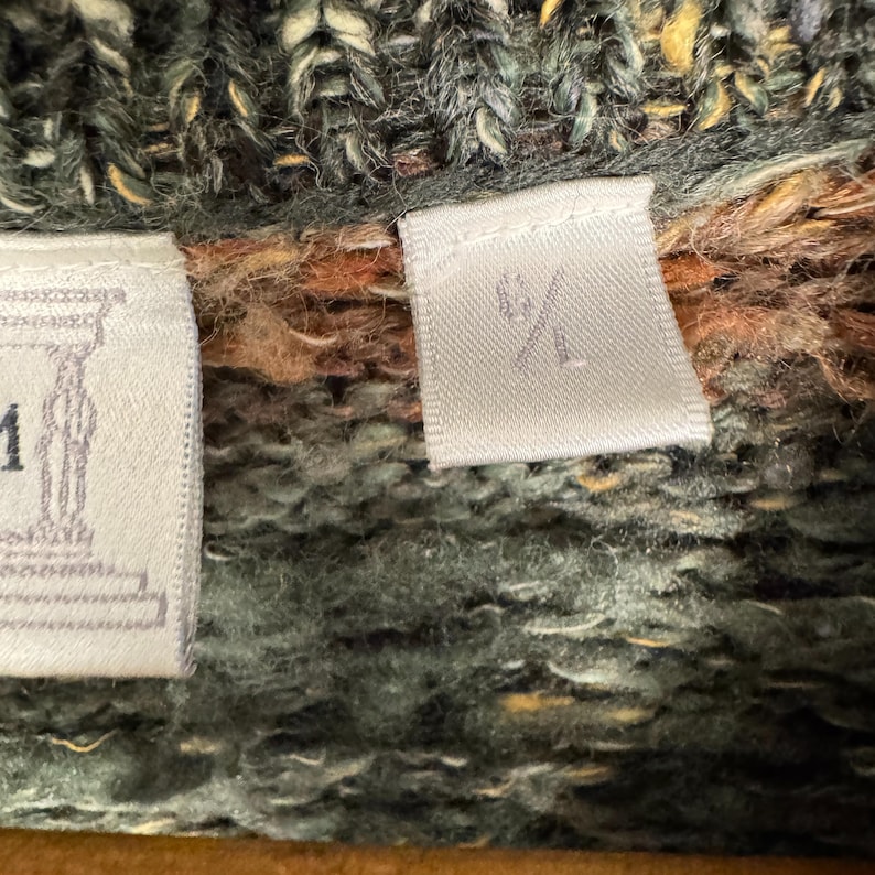 May include: Close-up of a textured knit sweater in shades of green, brown, and yellow. The sweater has a tag with the size marking "S/L". The knitwear appears to be thick and warm.