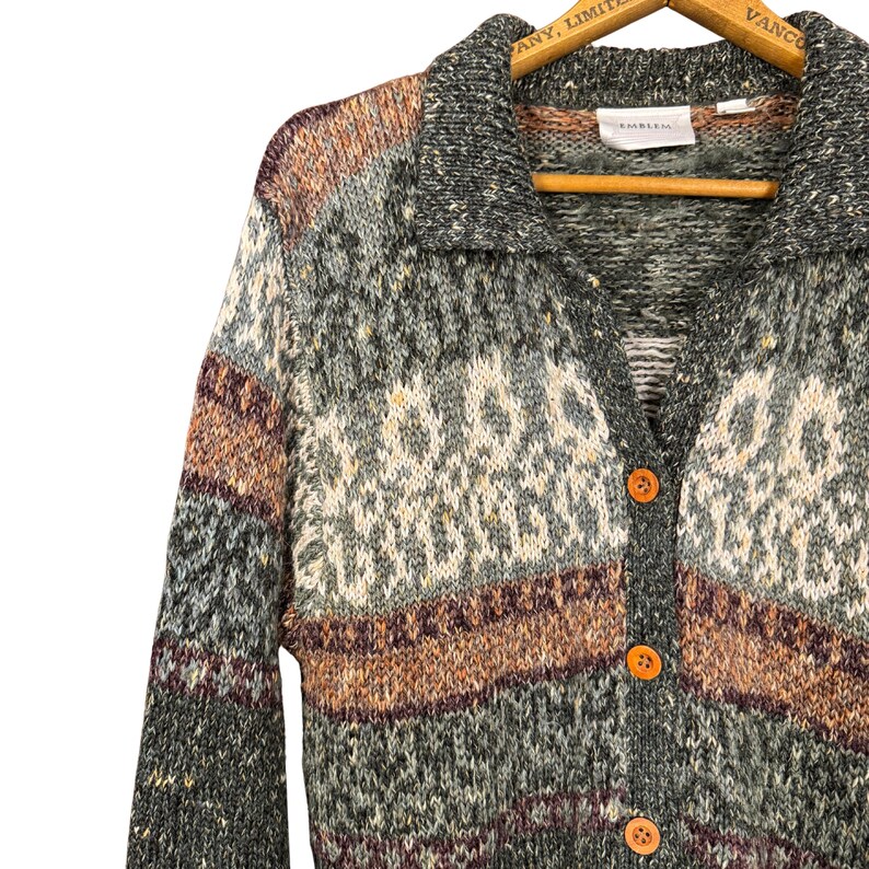 May include: A vintage button-up cardigan sweater with a collared neckline. The sweater features a textured knit pattern in shades of gray, green, brown, and cream. It has three orange buttons down the front and is hanging on a wooden hanger.