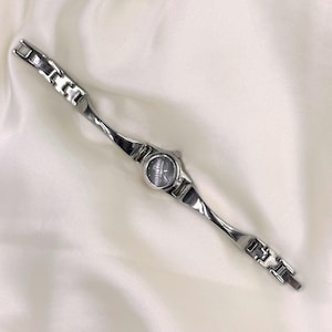 May include: A silver-toned wristwatch with a dark gray face and a linked band. The watch has a twisted design on the band, and the face has silver-toned hands and markers. The watch is displayed on a white, silky surface.