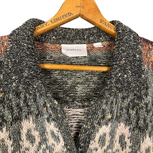 May include: A multi-colored knit cardigan with a collared neckline, hanging on a wooden hanger. The sweater features a mix of gray, green, and cream colors with a geometric pattern. The label reads "EMBLEM".