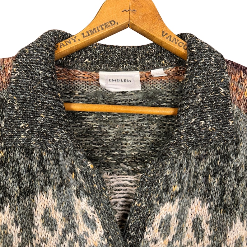 May include: A multi-colored knit cardigan with a collared neckline, hanging on a wooden hanger. The sweater features a mix of gray, green, and cream colors with a geometric pattern. The label reads "EMBLEM".