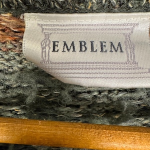 May include: Close-up of a woven label with the word "EMBLEM" in a serif font. The label is white with a light gray border and a decorative design. The background is a textured, green and brown knit fabric.