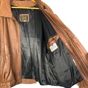 May include: A brown leather jacket with a black interior lining. The jacket has a zipper closure and a collared neckline. The interior features a pocket with a label. The jacket is open, revealing the lining and interior details.
