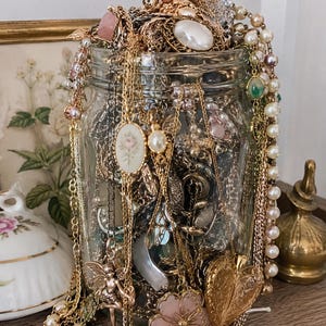 Peut inclure: Un bocal en verre transparent rempli d'un assortiment de colliers dorés, de rangs de perles et de divers pendentifs. On distingue un médaillon en forme de cœur, un camée floral, une breloque fée et un pendentif en forme de fleur rose.
