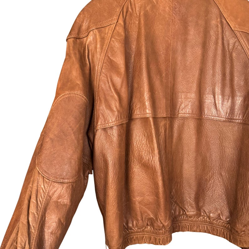 May include: A brown leather jacket with a classic design. The jacket features a rounded collar, shoulder pads, and a ribbed hem. The leather has a slightly distressed appearance, adding to its vintage appeal. The jacket is a medium brown color.