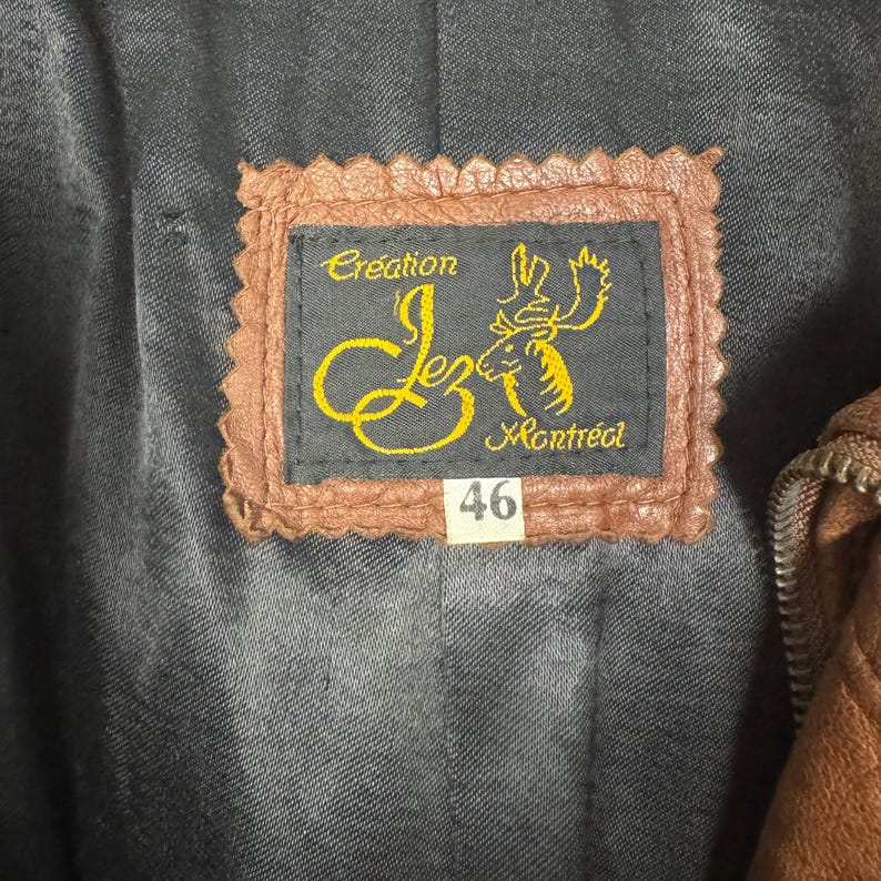 May include: Close-up of a brown leather label with the text "Creation Lez Montr&eacute;al" in gold lettering. The label also features a stylized deer graphic and the number "46" in a white box. The label is sewn onto a dark gray fabric.