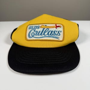 May include: A yellow and black baseball cap featuring a rectangular patch on the front. The patch displays the text Olds Cutlass in blue script lettering, accompanied by an embroidered graphic of a gold sword with a red handle.