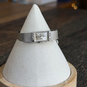 May include: A vintage silver wristwatch with a rectangular face and a mesh band. The watch face is white with black numerals and hands. Small diamonds are set on the watch face and band. The watch is displayed on a white cone-shaped stand.