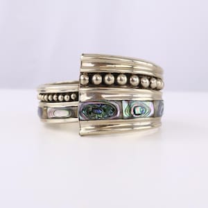 May include: A silver-toned cuff bracelet featuring a wide band with a polished finish. The design includes two rows of small, spherical metal beads and a central horizontal inlay of iridescent abalone shell pieces with green, blue, and purple hues.