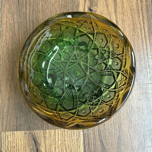 May include: A round, green and amber-colored glass bowl with a detailed starburst pattern. The bowl has a thick rim and a textured interior, creating a vintage aesthetic. The bowl is likely used for decorative purposes.