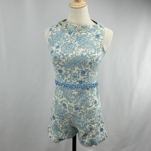 Puede incluir: Mono vintage con estampado de cachemira azul claro y crema. El mono sin mangas presenta un escote con volantes y un cinturón azul decorativo. Los pantalones cortos tienen un ligero vuelo. La prenda se exhibe en un maniquí.