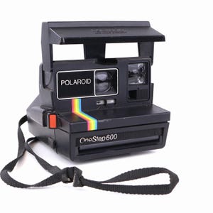 May include: A black Polaroid OneStep 600 instant camera featuring a rectangular body, a folding flash cover, and a prominent rainbow stripe graphic on the front. The device includes a black carrying strap and clearly displays the brand name.