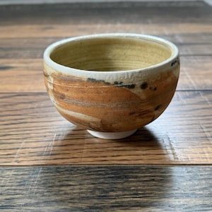 May include: A handmade ceramic bowl with a rounded shape. The bowl features a white base, a light brown interior, and a textured exterior with brown and black brushstrokes. The rim is white with small black markings.