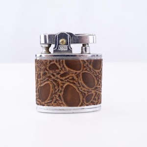 May include: A rectangular metal lighter featuring a brown textured wrap with an embossed pattern resembling reptile skin. The top mechanism is silver-toned metal with a small screw and the letters MC clearly visible on the front face.