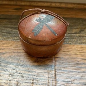 May include: A round, lidded ceramic box with a dragonfly design. The box is primarily reddish-brown with a darker brown dragonfly illustration on the lid. A brown cord handle is attached. The box is sitting on a wooden surface.