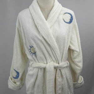 May include: Cream-colored terry cloth robe with a shawl collar and a matching belt. The robe features embroidered celestial designs: a sun, a crescent moon, and a moon with a face. The robe is displayed on a mannequin.