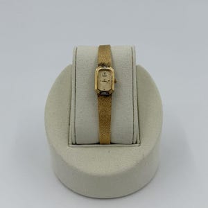 Puede incluir: Un reloj de pulsera dorado con una esfera rectangular y una correa a juego. El reloj se exhibe sobre un soporte color crema sobre un fondo blanco. La esfera del reloj tiene pequeños marcadores y agujas.