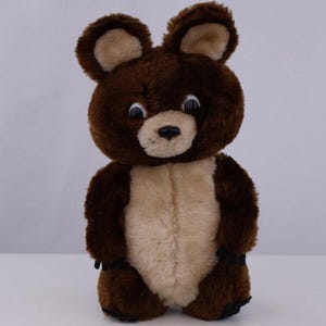 May include: A plush bear toy featuring dark brown fur on the head, ears, and limbs, with a contrasting light beige patch on the chest and face. The bear has large, round, dark eyes with white pupils and a small black nose, standing in an upright position.