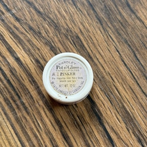 Op de afbeelding: Een vintage Yardley Pot o' Gloss getinte lipgloss in een kleine, ronde verpakking. Het etiket luidt "Pinker" en "Net Wt. 12 oz." De verpakking is wit met een gouden rand en tekst. De achtergrond is een houten oppervlak.