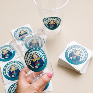 May include: Clear plastic cups and circular stickers with a teal and white checkered border. The stickers and cups feature a photo of a person and the text "Justin's 30th Birthday".