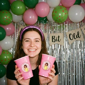 May include: A smiling person wearing a glitter headband holds two pink cups with a circular sticker that says "Look Who's 30". The background includes balloons in pink, green, and white, and a banner that says "Bit Old".