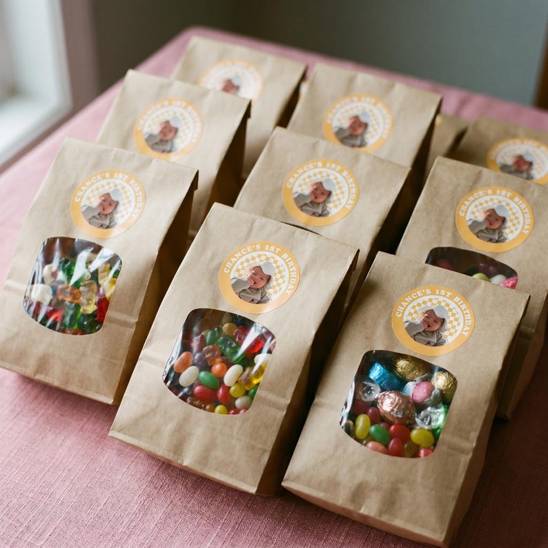 May include: Brown paper treat bags with clear windows filled with colorful candies. Each bag has a circular sticker that reads "Chance's 1st Birthday" with a baby's photo. The bags are arranged on a pink surface.