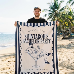 May include: A rectangular beach towel featuring a cream-colored center with dark blue text reading WELCOME TO SHINTAROH'S BACHELOR PARTY and BALI ISLAND. The graphic shows a person in a suit sitting in a martini glass. The border has blue stripes.