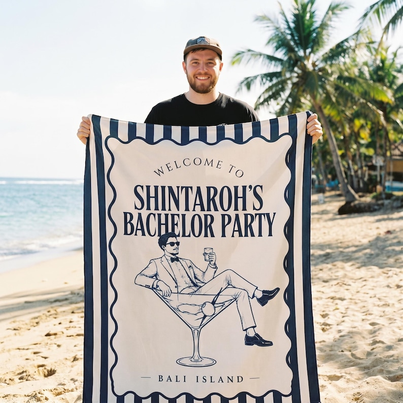 May include: A rectangular beach towel featuring a cream-colored center with dark blue text reading WELCOME TO SHINTAROH'S BACHELOR PARTY and BALI ISLAND. The graphic shows a person in a suit sitting in a martini glass. The border has blue stripes.