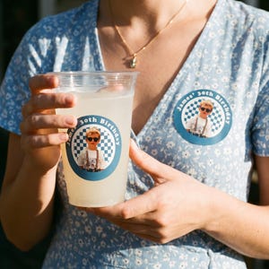 May include: A clear plastic cup filled with a light yellow liquid, featuring a circular sticker that reads "James' 30th Birthday" with a photo of a child wearing sunglasses. A matching sticker is on the blue floral dress.