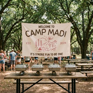May include: A rectangular off-white fabric banner featuring the brown text 'WELCOME TO CAMP MADI', 'EST. 2024', and 'IT'S S'MORE FUN TO BE ONE', with a red line-art illustration of mountains, trees, a tent, and a sun centered on the banner.