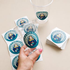 May include: Clear plastic cups and circular stickers with a teal and white checkered border. The stickers and cups feature a photo of a person and the text "Justin's 30th Birthday".