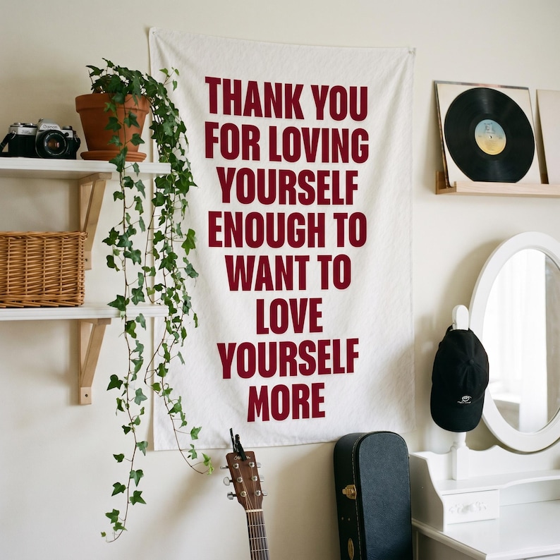 May include: A white fabric wall hanging featuring the dark red text: THANK YOU FOR LOVING YOURSELF ENOUGH TO WANT TO LOVE YOURSELF MORE. The banner is rectangular with a clean, simple font and is displayed vertically against a plain wall.