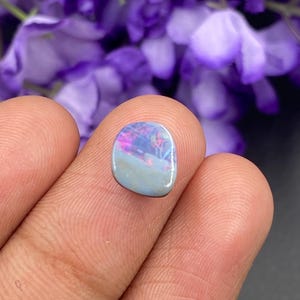 Puede incluir: Una piedra preciosa de ópalo pulida, de forma ovalada. El ópalo presenta una base azul claro con una vibrante exhibición de colores internos rosas, morados y azules. La piedra preciosa se sostiene entre dos dedos, mostrando su tamaño y detalle.