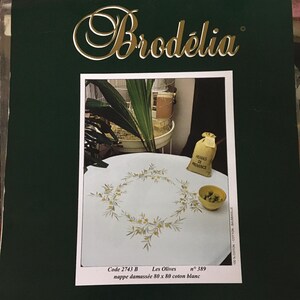 May include: A white tablecloth with a green olive branch embroidery design. The tablecloth is labeled "Les Olives" and is part of the Brodelia collection by Margot Creations of Paris. The tablecloth is 80 x 80 centimeters.