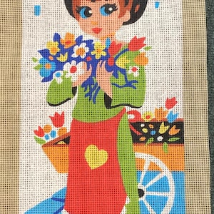 May include: Needlepoint canvas depicting a girl with flowers in front of a Parisian scene. The girl is dressed in green with a red apron and a yellow heart. The background shows buildings and a flower cart. The text "Royal Paris" is visible.