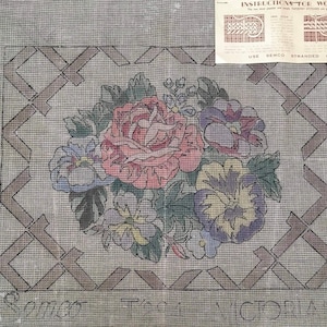May include: A needlepoint canvas with a floral design featuring a pink rose, purple pansies, and green leaves. The canvas is surrounded by a brown lattice pattern. The text "VICTORIA" is visible at the bottom of the canvas.